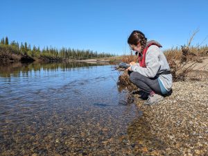 A woman holding, and looking at, a device crouches down next to a small creek with a rugged shoreline in the background.
