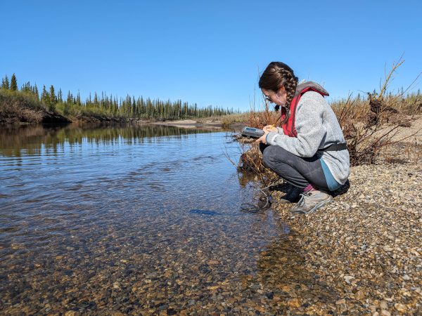 A woman holding, and looking at, a device crouches down next to a small creek with a rugged shoreline in the background.