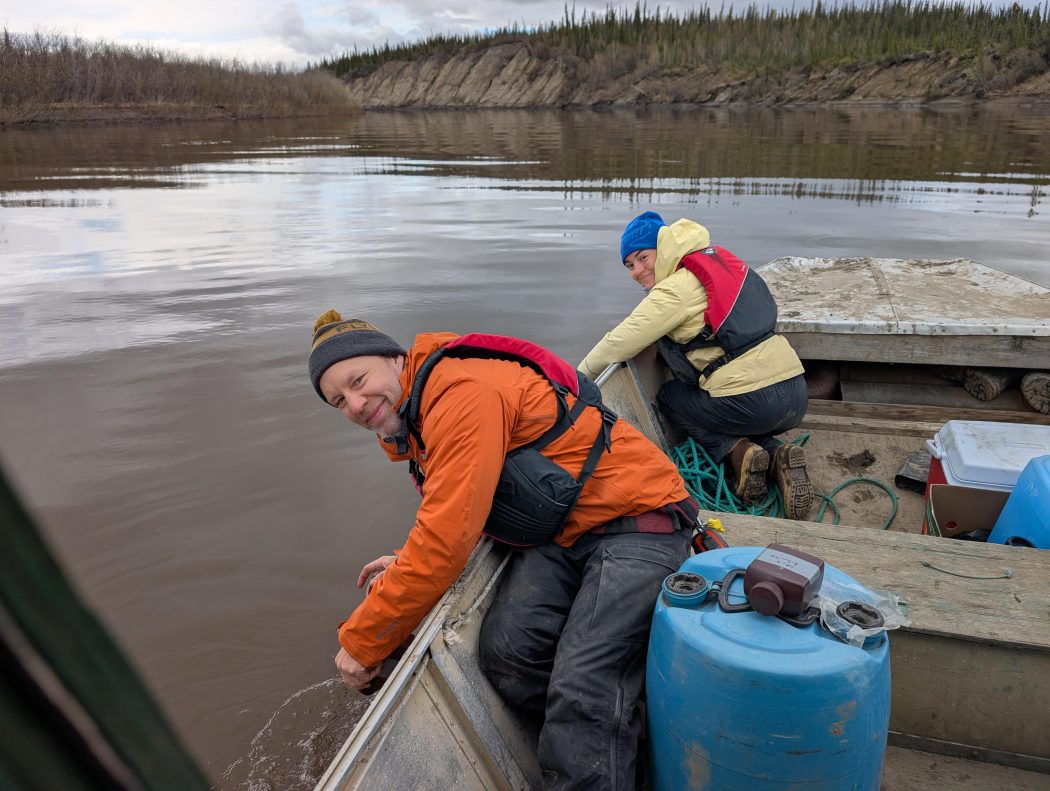 A man and a woman are crouching and leaning over the side of a boat, holding a net into the water, with a rocky shoreline in the background.