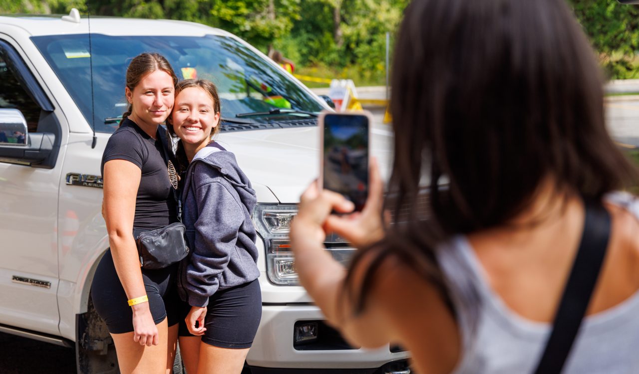 A woman takes a photo of two young women standing beside a car.