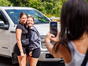 A woman takes a photo of two young women standing beside a car.