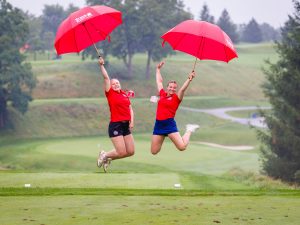 Two people in red shirts jump in the air with red umbrellas on a golf course.