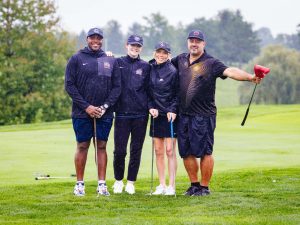 Four people pose with golf clubs on a golf course.