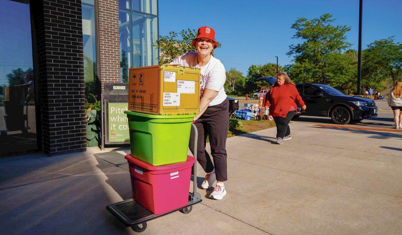 A woman in a red bucket had with a large "B" on it smiles as she moves two large plastic totes and a box on a trolley into a building.