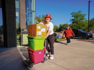 A woman in a red bucket had with a large "B" on it smiles as she moves two large plastic totes and a box on a trolley into a building.