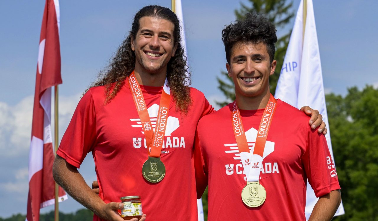 Two men stand against a blue sky wearing red T-shirts and gold medals hanging around their necks.