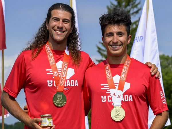 Two men stand against a blue sky wearing red T-shirts and gold medals hanging around their necks.