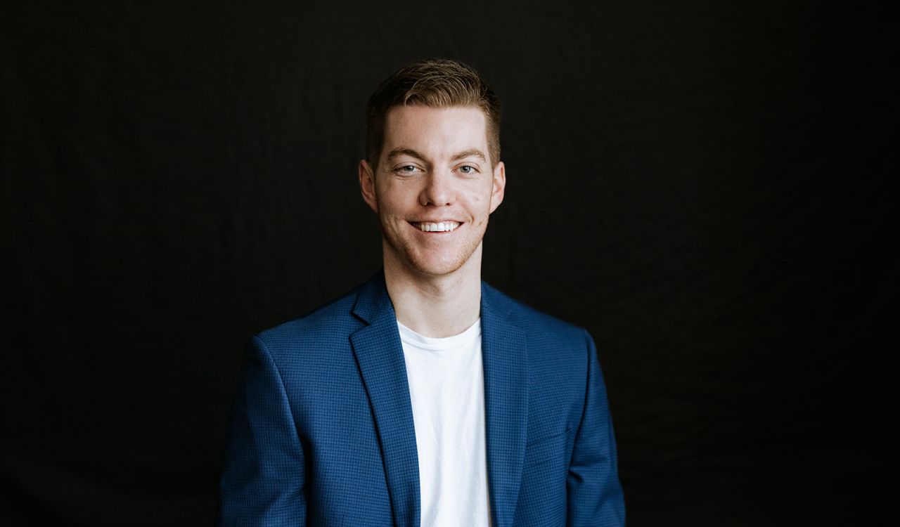 Brad Rossen stares straight ahead, smiling, with a black textured background behind him.