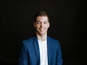 Brad Rossen stares straight ahead, smiling, with a black textured background behind him.