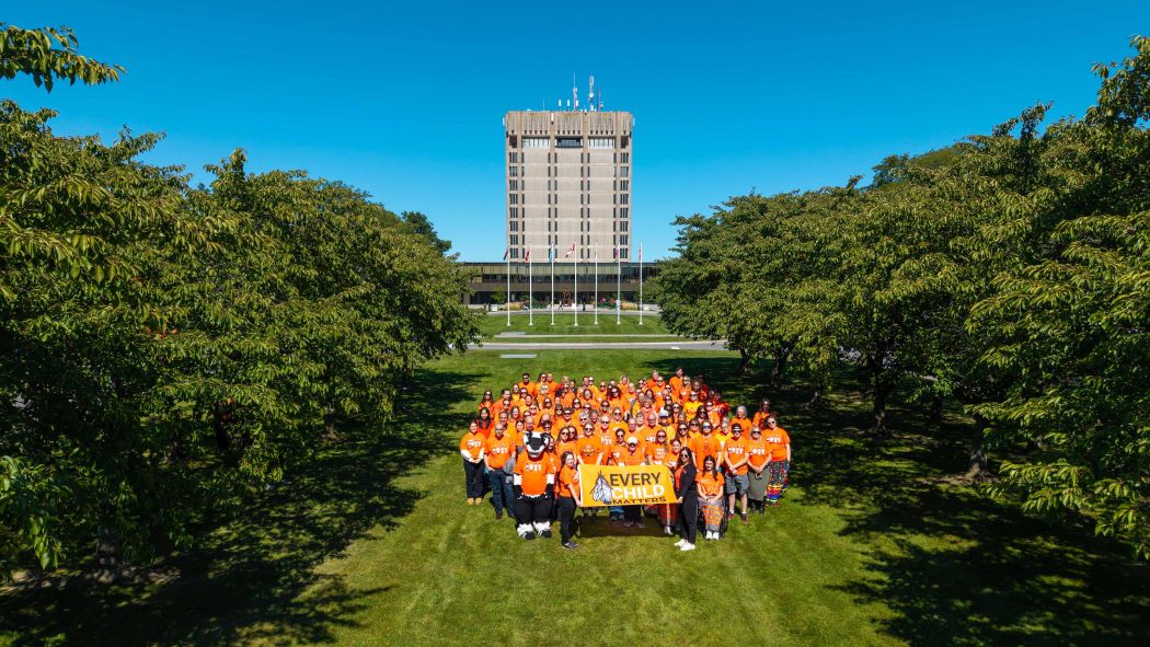 A large group of people in orange shirts pose with an Every Child Matters flag in front of Brock University's Schmon Tower.