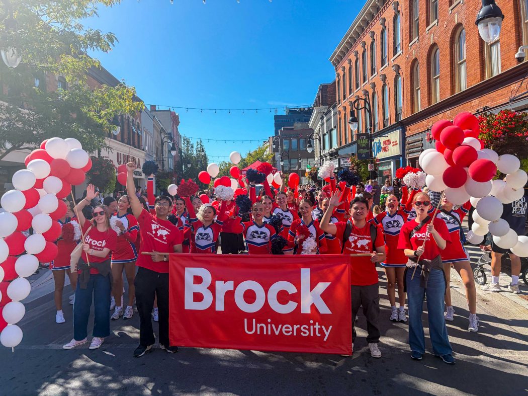 A large group of people in red shirts and cheerleading uniforms walk behind a Brock University banner at a parade.