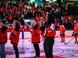 A woman sings Canada's national anthem into a microphone while standing on the ice in a packed hockey arena.
