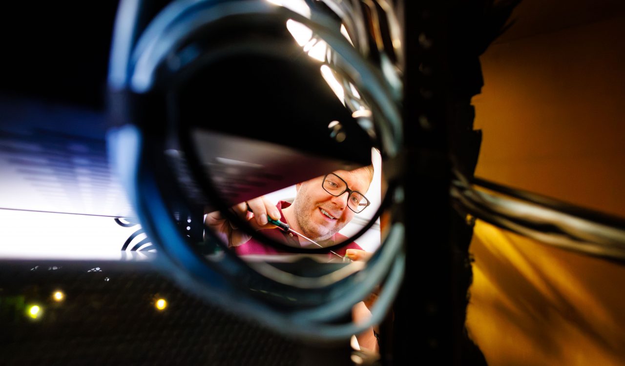 Greg Beaudry from the audio-visual team working inside a network equipment rack, viewed through a loop of blue cables while using a screwdriver to connect or adjust cabling.