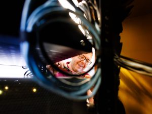 Greg Beaudry from the audio-visual team working inside a network equipment rack, viewed through a loop of blue cables while using a screwdriver to connect or adjust cabling.