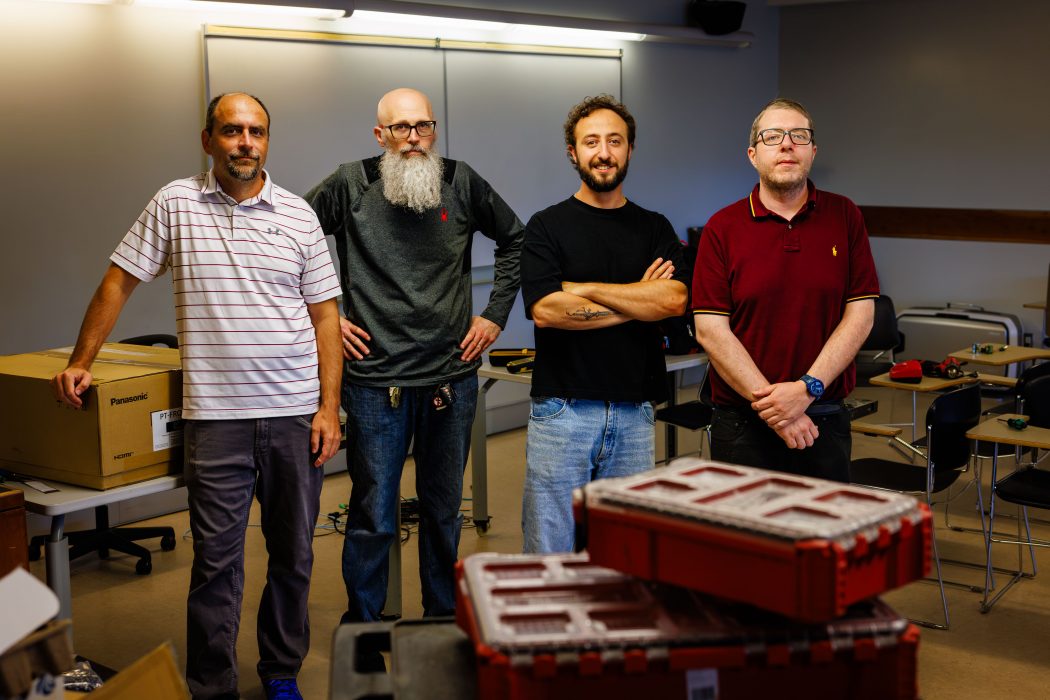 Milos Maslek, Gary Cigic, Can Kilic and Greg Beaudry stand together in a classroom space during the AV modernization project. Equipment boxes, protective cases and classroom furnishings are visible around them.
