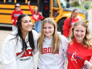 Three women in Brock University shirts stand outside in front of a school bus.