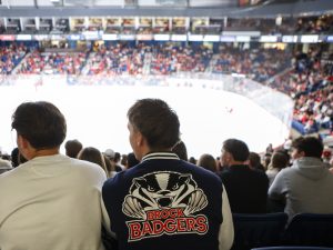 A packed arena watches a hockey game. A man sits with a Brock Badgers varsity jacket on.