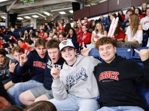 A group of men in Brock University shirts sit in a packed hockey arena.