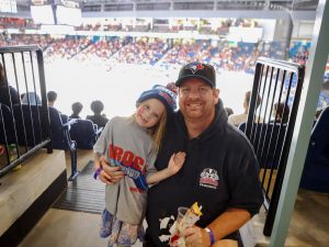 A child with Brock Badgers face painting and a Brock t-shirt stands with a man in a Brock Badgers sweatshirt in front of a hockey arena full of people.