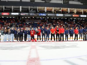 A large group of people stands on the ice of a hockey arena with two trophies in hand.