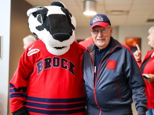 A man in a Brock University hat and jacket stands next to Boomer the Badger, Brock University's mascot.