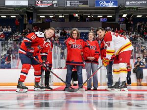 The captains of the Brock Badgers and Guelph Gryphons face off with their sticks to the ice as Brock University President Lesley Rigg drops the puck at centre ice in a packed hockey arena.