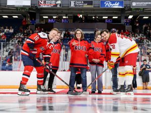 The captains of the Brock Badgers and Guelph Gryphons face off with their sticks to the ice as Brock University President Lesley Rigg drops the puck at centre ice in a packed hockey arena.