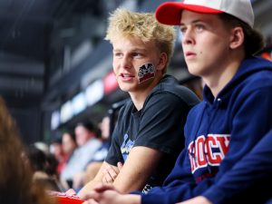 Two men sit in stadium seats, one with a large Brock Badgers sticker on his cheek and the other wearing a Brock University sweatshirt.