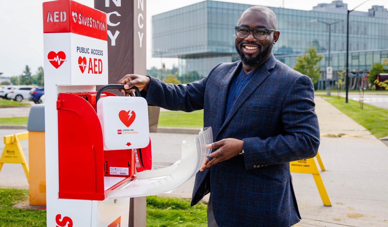 Director of Health, Safety and Wellness James Iseghayan stands beside an outdoor automated external defibrillator (AED) station on Brock University’s main campus, opening the cabinet and holding an AED. A campus building is visible in the background.