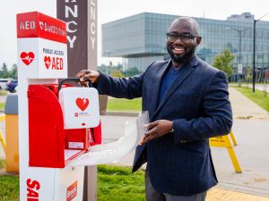 Director of Health, Safety and Wellness James Iseghayan stands beside an outdoor automated external defibrillator (AED) station on Brock University’s main campus, opening the cabinet and holding an AED. A campus building is visible in the background.