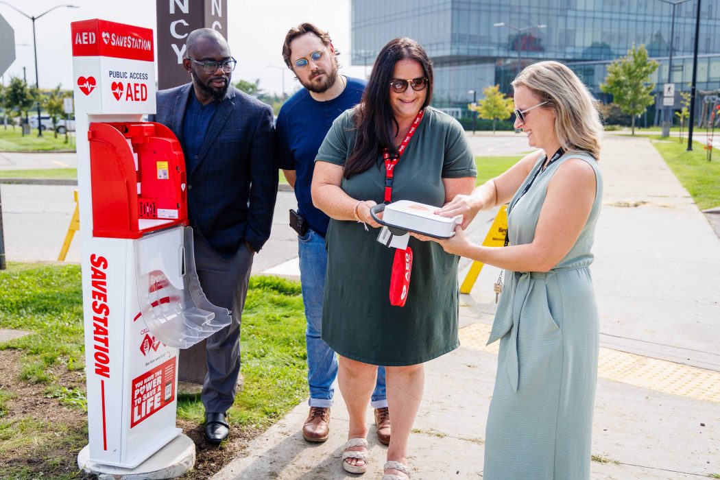 James Iseghayan, Evan Schipper, Elizabeth Todd and Britton Sheppard examine an automated external defibrillator that has been removed from the cabinet. A modern glass building and landscaped grounds are visible in the background.