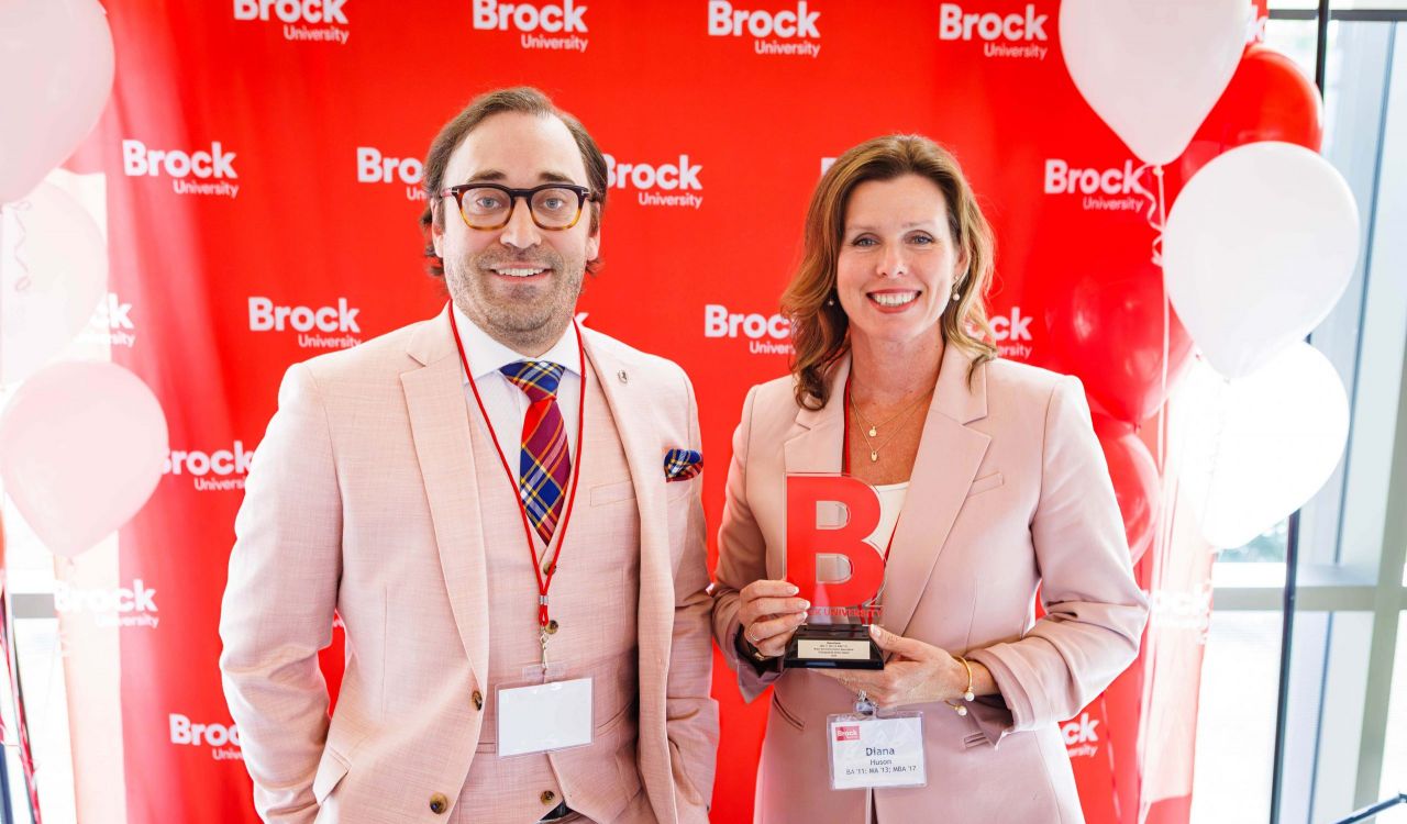 A man and a woman stand in front of a red backdrop. The woman is holding an award.