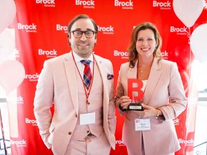 A man and a woman stand in front of a red backdrop. The woman is holding an award.