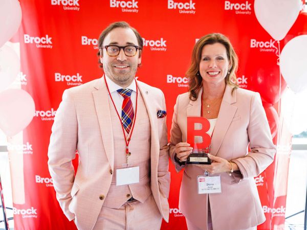 A man and a woman stand in front of a red backdrop. The woman is holding an award.