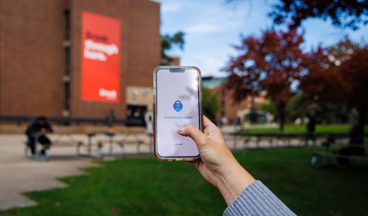 A hand holds a smartphone displaying the Microsoft Authenticator app on Brock University's campus, with a campus building and Brock banner blurred in the background.