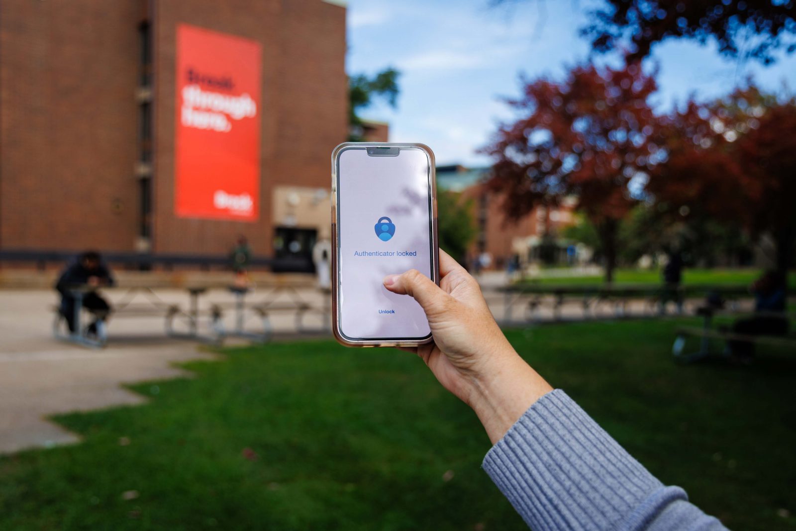 A hand holds a smartphone displaying the Microsoft Authenticator app on Brock University's campus, with a campus building and Brock banner blurred in the background.