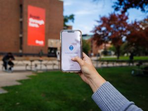 A hand holds a smartphone displaying the Microsoft Authenticator app on Brock University's campus, with a campus building and Brock banner blurred in the background.