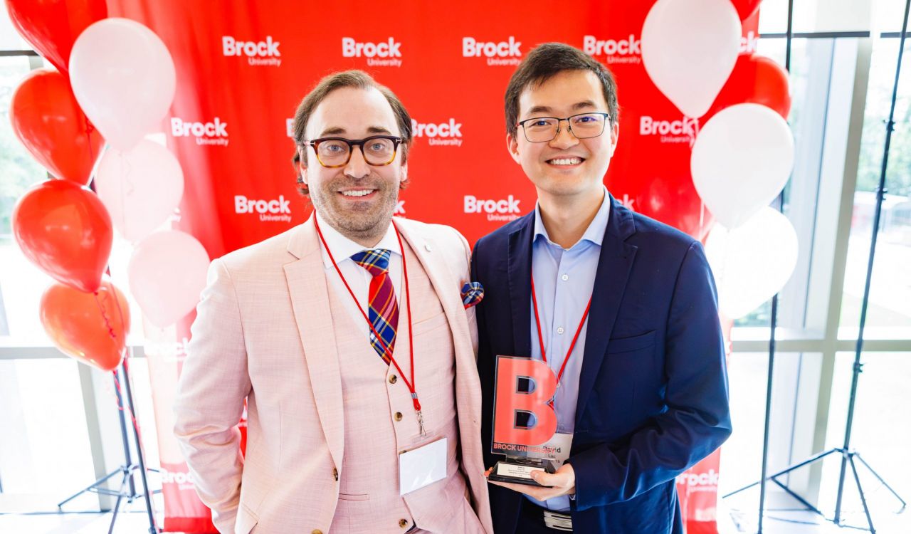 Two men stand in front of a red Brock University backdrop, with one holding an award.