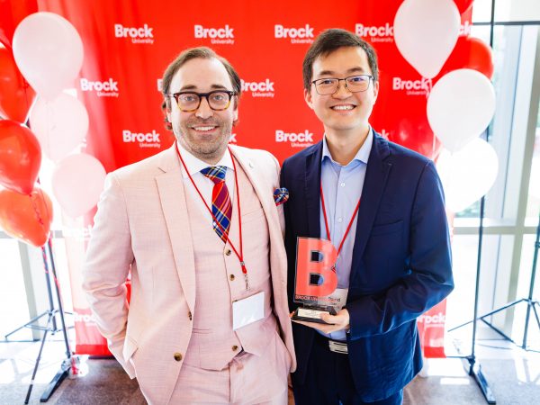 Two men stand in front of a red Brock University backdrop, with one holding an award.