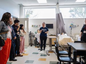 A woman in a nursing simulation lab give a group a tour. There are mannequins in hospital beds in the room.