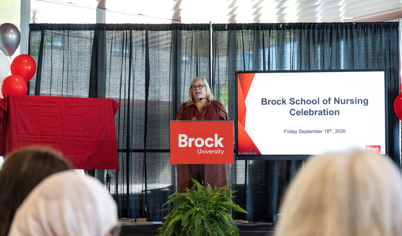 Karyn Taplay, Director and Associate Professor of Brock's School of Nursing speaks into a microphone at a podium while addressing a room of people.