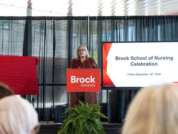 Karyn Taplay, Director and Associate Professor of Brock's School of Nursing speaks into a microphone at a podium while addressing a room of people.