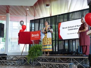 A woman beats a hand drum while standing on a stage at a Nursing Celebration event.
