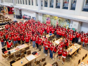 A large number of students in red shirts pose for a group photo in a university cafeteria.