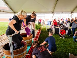Two women stomp grapes in barrels in front of guests at a wine industry event.