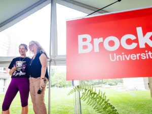 Two smiling women standing under a tent beside a Brock University banner, one holding a small trophy and wearing a ‘grapes’ T-shirt.
