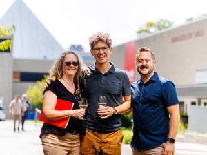 Three people smiling together outdoors on campus, two holding wine glasses, with a red Brock University banner in the background.