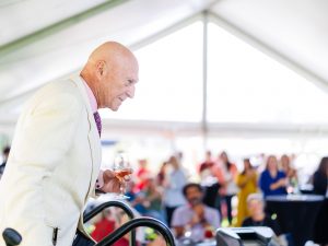 An older man in a cream blazer and pink shirt holding a wine glass, looking out over a crowd seated under a large event tent.