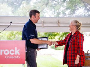 Mayor of St. Catharines shaking hands with a woman in a red plaid blazer under a tent, presenting a framed certificate near a Brock University sign.