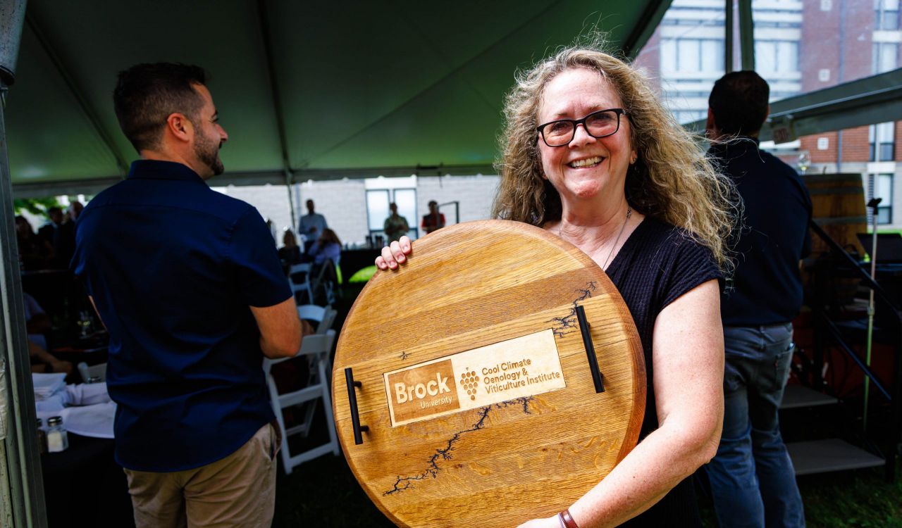 Barbara Tatarnic holds a large wooden display made from a wine barrel head featuring the Brock University Cool Climate Oenology and Viticulture Institute logo while standing under a tent at an outdoor event.